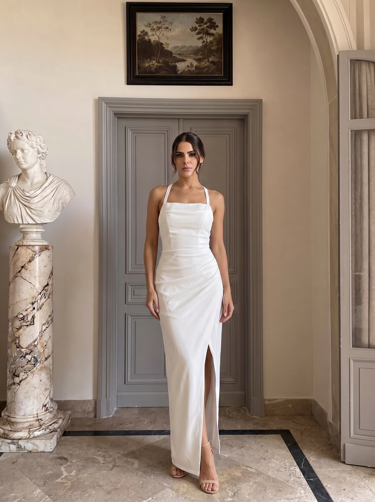 A woman stands indoors wearing a sleeveless white maxi dress with thin straps, a fitted ruched bodice, and a high front slit. The setting includes a gray paneled door, a marble bust on a column, and a framed landscape painting on the wall.