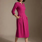 Woman wearing a diva catwalk Amanda Pleated a-line dress in a magenta pink colour with pockets against a beige background (Magenta)