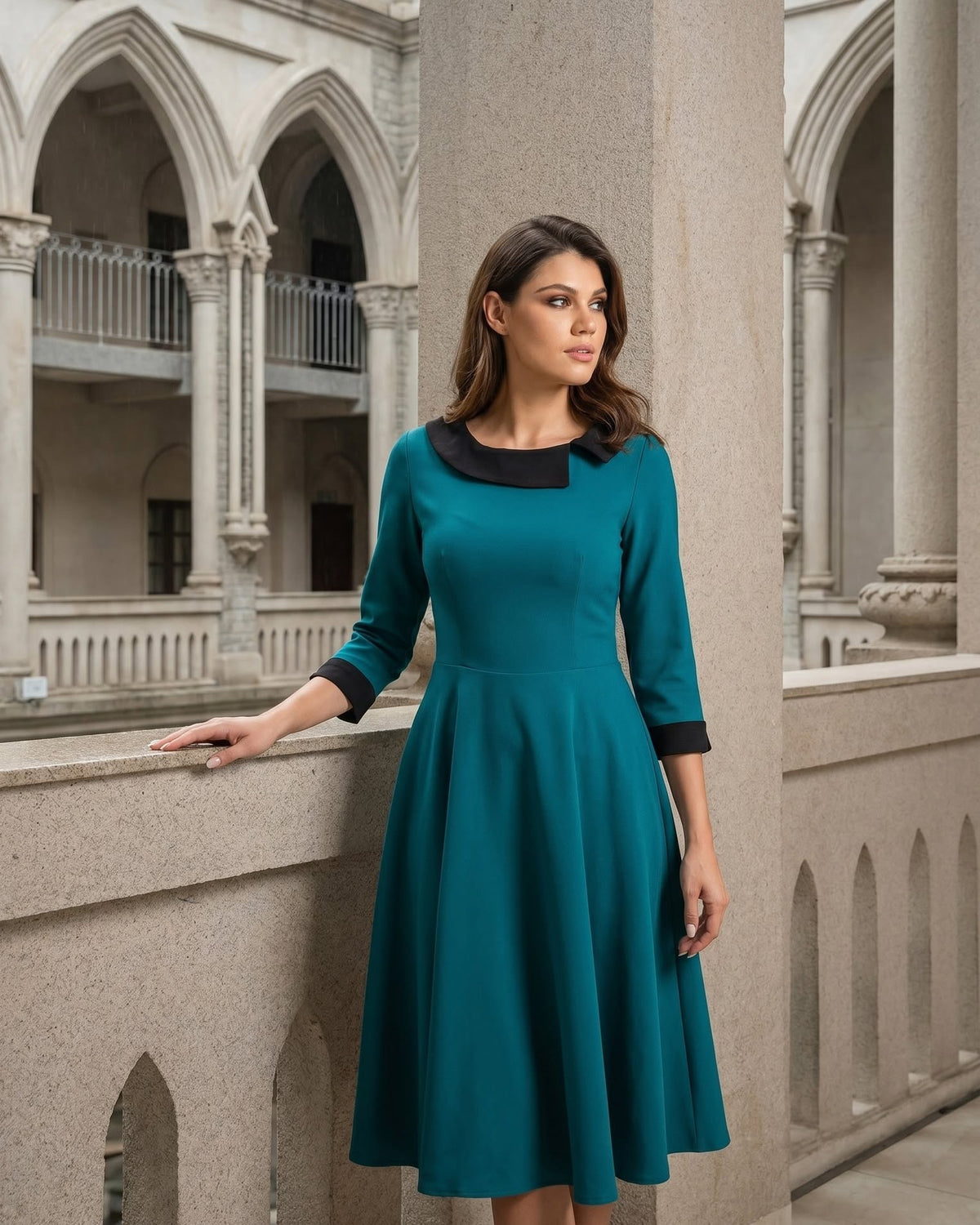 A woman wearing a teal three-quarter sleeve dress with a black collar and cuffs, standing in a stone architectural setting with arches.
