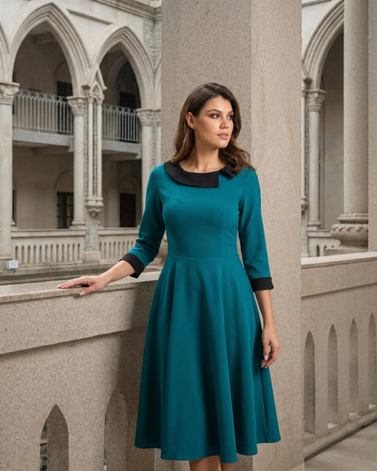 A woman wearing a teal three-quarter sleeve dress with a black collar and cuffs, standing in a stone architectural setting with arches.